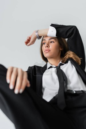 Foto de Young brunette woman in black formal wear with tie looking away while posing on grey background - Imagen libre de derechos