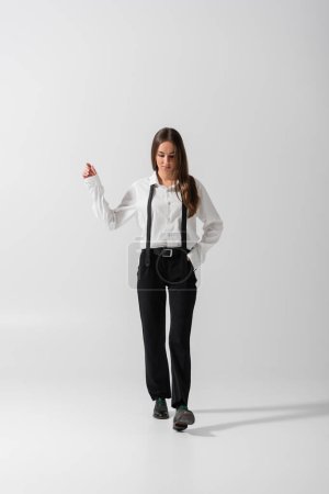 Foto de Full length of brunette woman in black pants and suspenders walking with hand in pocket on grey - Imagen libre de derechos