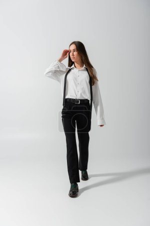 Photo for Full length of brunette young woman in black pants and suspenders walking on grey - Royalty Free Image