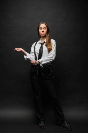 full length of brunette woman in formal wear with suspenders standing and looking away on black 