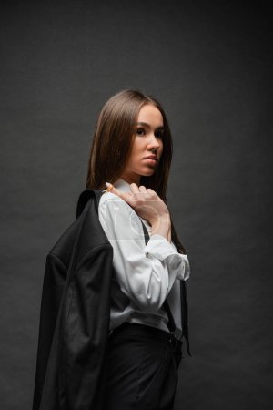 brunette woman with long hair standing in formal wear and holding blazer on black 