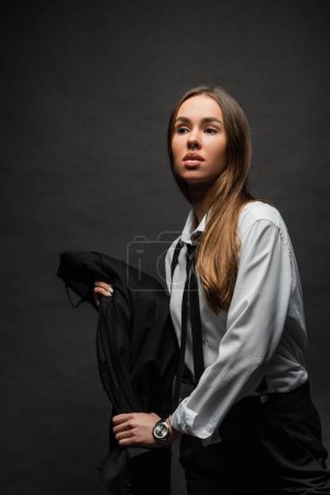 young brunette woman with long hair standing in suit while holding blazer on black background 