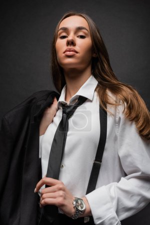 low angle view of confident woman in suit holding jacket and looking at camera on black 