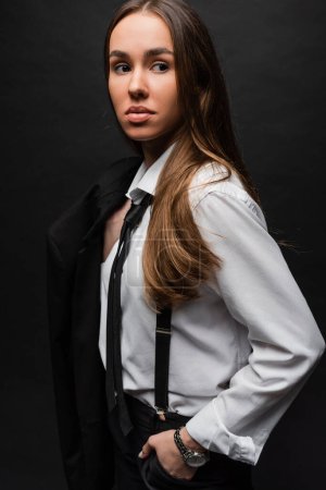 young woman with brunette long hair standing in suit and holding blazer on black 