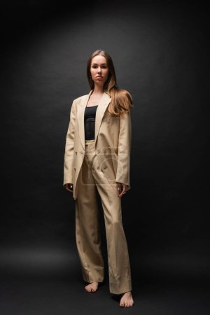 full length of barefoot woman in beige suit standing and looking at camera on black 