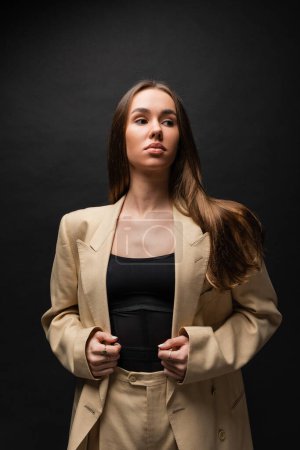 brunette young woman in beige blazer standing and looking away on black 