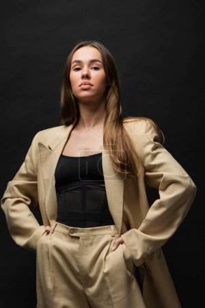 brunette young woman in beige suit standing with hands in pockets on black 