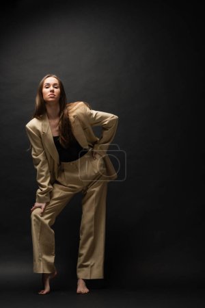 full length of barefoot young woman in beige suit posing with hand on hip on black 