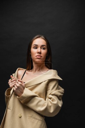 brunette young woman adjusting beige blazer and looking away on black 