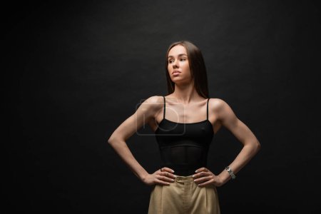 Photo for Brunette and muscular woman in black top and beige pants posing with hands on hips on black background - Royalty Free Image