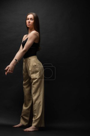 full length of barefoot woman in top and beige pants standing with clenched hands on black background 