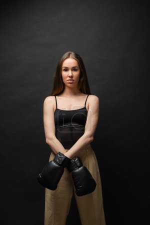 brunette woman in top and beige pants posing in boxing gloves on black background 