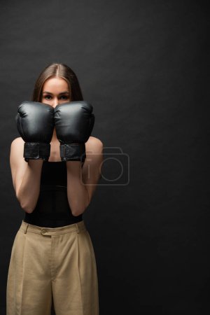 brunette and strong young woman in top covering face with boxing gloves on black background 