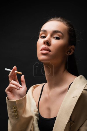 young brunette woman in beige blazer holding cigarette while smoking on black 