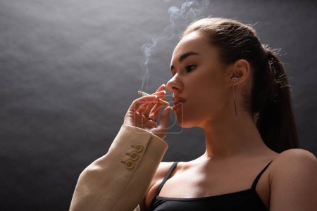 low angle view of young brunette woman in beige blazer smoking cigarette on black background 