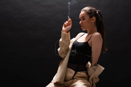 brunette woman in beige suit holding cigarette and sitting on black background 