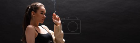 Foto de Young brunette woman in beige blazer holding cigarette while looking away on black background, banner - Imagen libre de derechos