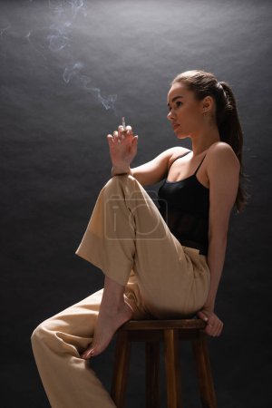 Barfüßige Frau in beiger Hose mit Zigarette auf dem Hochstuhl vor schwarzem Hintergrund 