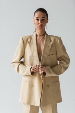 brunette model adjusting beige jacket posing while looking at camera isolated on grey 