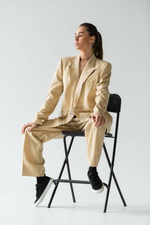 Brünette junge Frau im beigen trendigen Anzug sitzt auf Stuhl auf grau 