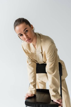 Foto de Pretty young woman in beige jacket leaning on chair back while looking at camera isolated on grey - Imagen libre de derechos