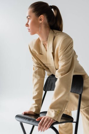 Foto de Pretty young woman in beige jacket leaning on chair while looking away isolated on grey - Imagen libre de derechos