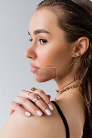 portrait of brunette woman in black top looking away while touching shoulder isolated on grey 