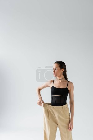 pretty model in black top looking away and adjusting oversized pants on grey 