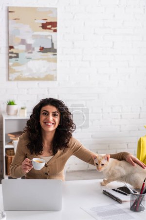 Téléchargez les photos : Smiling freelancer holding coffee and petting oriental cat near devices at home - en image libre de droit