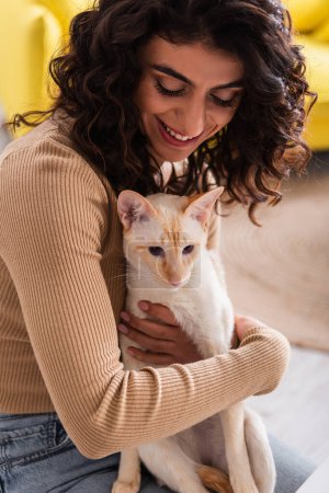 Smiling brunette woman hugging oriental cat at home 