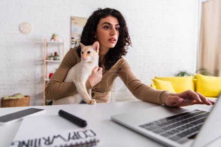 Copywriter holding oriental cat and using laptop near blurred notebook at home 
