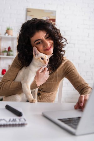 Positive woman holding oriental cat and using blurred laptop at home 