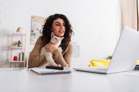 Smiling copywriter holding oriental cat and looking at laptop in living room 