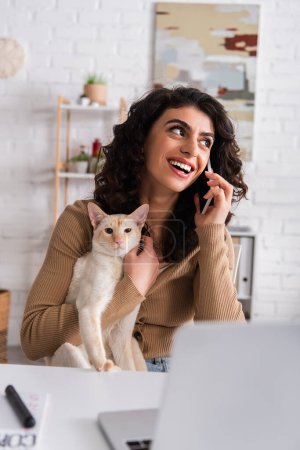 Smiling freelancer talking on smartphone and holding oriental cat near laptop at home 