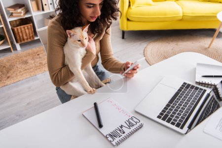 Rédactrice brune utilisant un smartphone et tenant un chat oriental à la maison 
