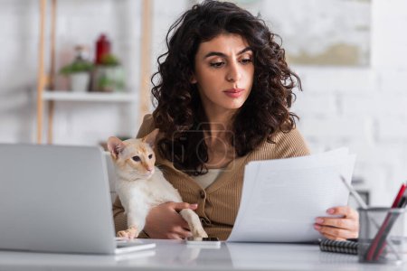 Morena redactor sosteniendo oriental gato y papeles cerca de portátil en casa 