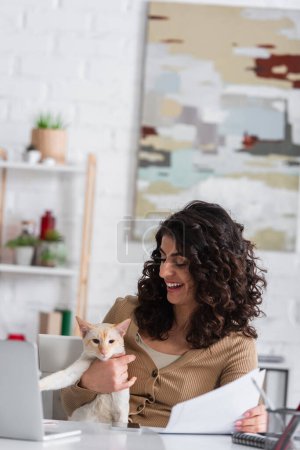 Téléchargez les photos : Smiling copywriter looking at oriental cat and holding papers near laptop at home - en image libre de droit