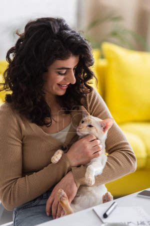 Smiling woman petting oriental cat near notebook and smartphone at home 