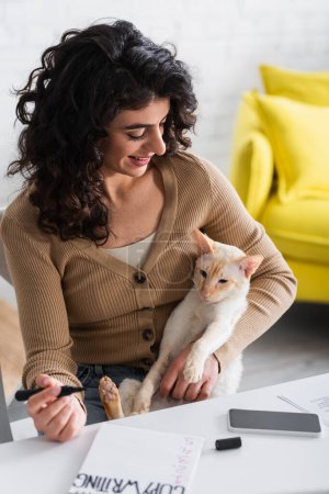 Foto de Redactor sonriente sosteniendo marcador y mirando a gato oriental cerca de portátil y teléfono inteligente - Imagen libre de derechos