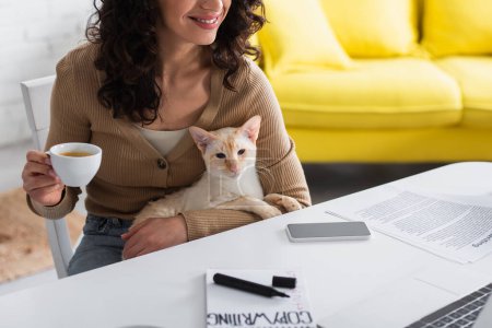 Foto de Vista recortada de un redactor sonriente sosteniendo una taza de café y un gato oriental cerca de dispositivos en casa - Imagen libre de derechos