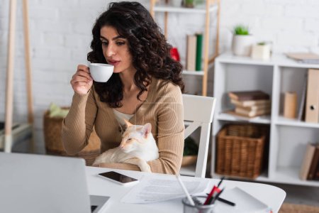 Brünette Werbetexterin hält Kaffee und orientalische Katze in der Nähe von Geräten und Papieren zu Hause 