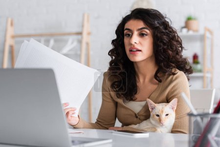 Brunette copywriter holding papers and oriental cat near laptop at home 
