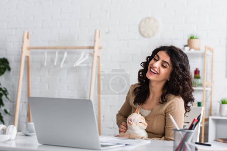 Cheerful copywriter holding oriental cat and looking at laptop near papers and headphones at home 