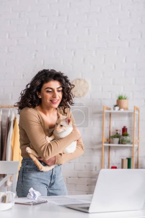 Positive freelancer holding oriental cat near laptop at home 