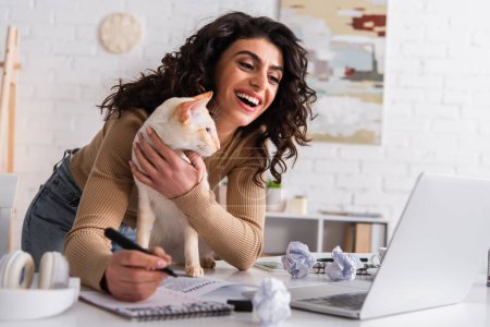 Heureux rédacteur caressant chat oriental près des papiers et ordinateur portable à la maison 