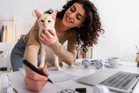 Brunette copywriter petting oriental cat near papers and laptop in living room 
