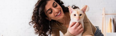 Smiling brunette woman looking at oriental cat at home, banner 