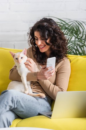 Lächelnder Werbetexter hält Handy und orientalische Katze neben Laptop auf Couch 