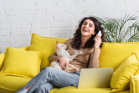 Alegre freelancer sosteniendo smartphone y gato oriental cerca de portátil en sofá 