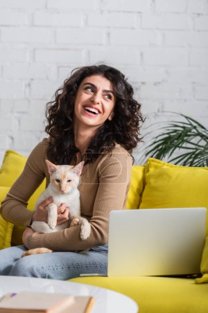 Smiling freelancer holding oriental cat near devices on couch 